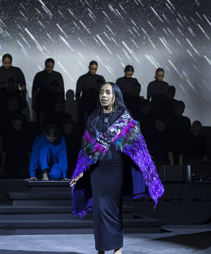 A performer in a vibrant Te Pae cloak stands barefoot, expressing emotion on stage. Behind, a group in black watches attentively, illuminated against a dramatic, rain-like backdrop.